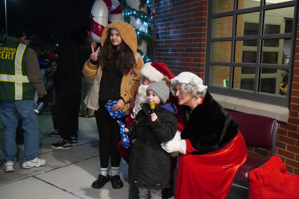 Santa Claus, Mrs. Claus with boy and girl pose for a photo. Girl gives a peace sign while the boy holds a bell.